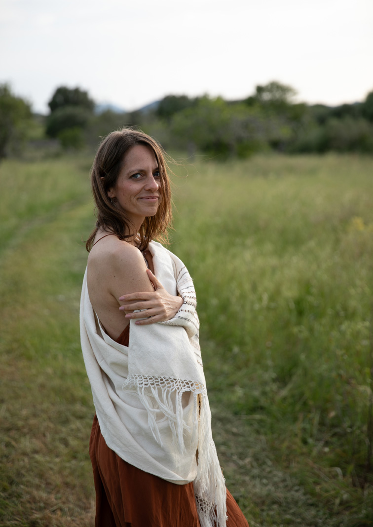 Waldpriesterin Katrin Sommer Eine Frau steht auf einem Weg in deiner Wiese, hat ein weißes schamanisches Tuch um sich gelegt und lächelt in die Kamera.