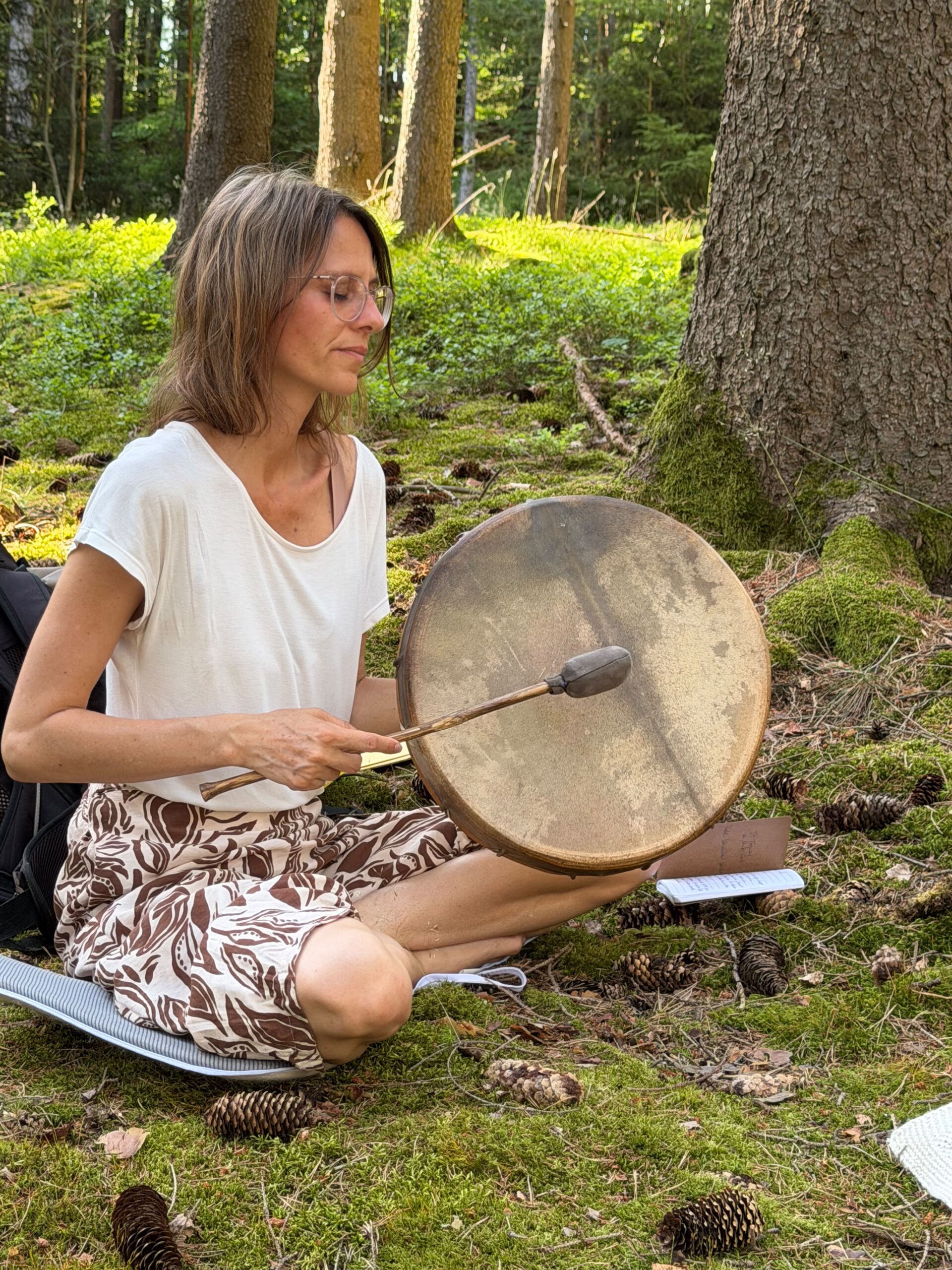 Trommeln mit der Urkraft von Mutter Erde Eine Frau trommelt auf einer schamanischen Trommel im Wald, sitzend in Meditationspose und geschlossenen Augen